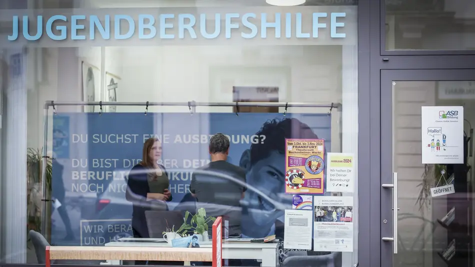 Blick durch das Schaufenster, das mit 'Jugendberufshilfe' beklebt ist: Zwei Menschen stehen im Gespräch in den Räumlichkeiten der Jugendberufshilfe Höchst.