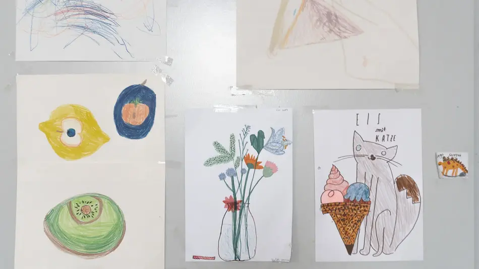 Fünf Kinderzeichnungen im betreuten Kinderzimmer der Hochschule für Gestaltung: Drei gut erkennbar. Links Obst, in der Mitte eine Vase mit Blumen, ganz rechts eine Katze mit Eis mit dem Titel "Eis mit Katze" zu sehen.